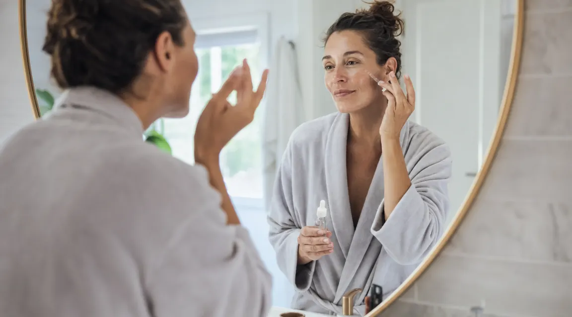 Woman applying serum to cheek in bathroom mirror, morning skincare routine